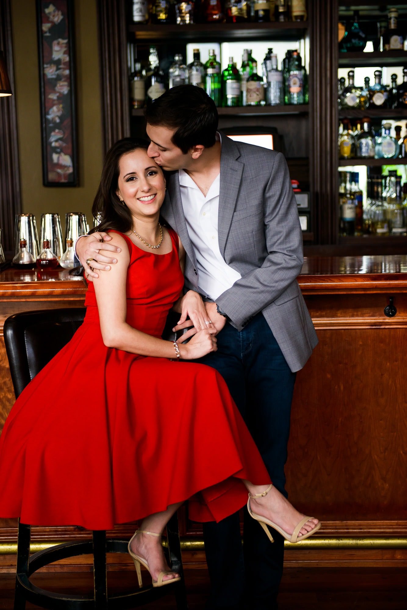 Vanessa & Matthew's Downtown San Antonio Engagement Session | Man embracing his fiancé and kissing her on the head at bar in Bohanan's. Red dress with nude heels, gray suit.
